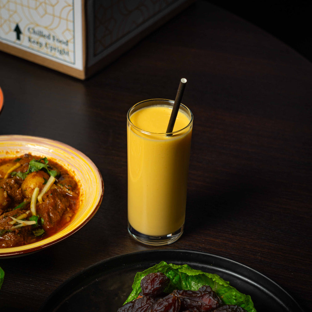 Glass of mango lassi with a straw on a dark surface with food items.