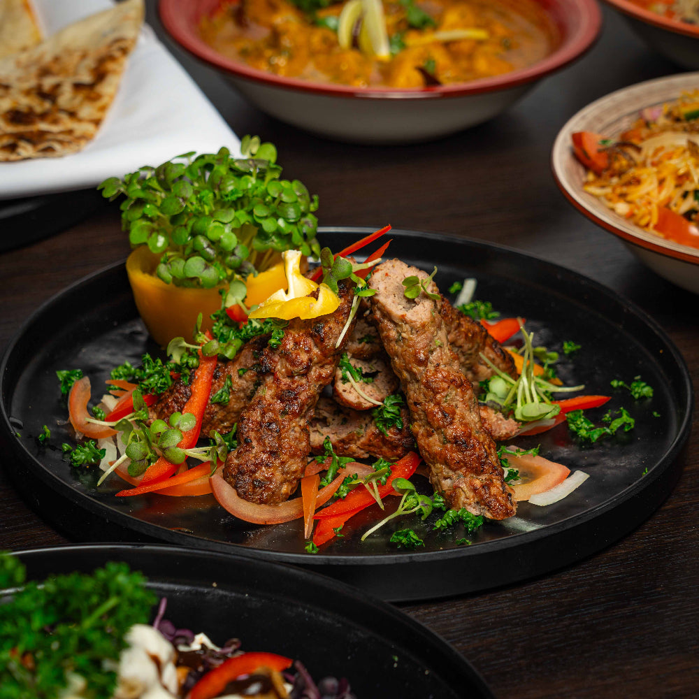 Plated dish of grilled meat with vegetables on a dark surface, surrounded by other dishes.