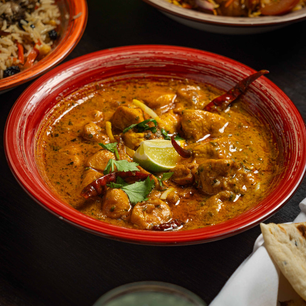 Red bowl of curry with chicken, lime, and chili peppers on a dark surface.